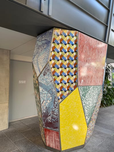 The image features a large, multi-faceted, polygonal sculpture or public seating element situated in South Brisbane, Australia. This striking object is constructed from numerous irregular panels, each adorned with a distinct pattern or texture.

Prominently displayed are panels with abstract designs: a red segment with a white swirling pattern, a green panel with a similar texture, and a bright yellow panel featuring a white mottled surface. A central section showcases a vibrant geometric pattern composed of interlocking rhombuses and triangles in hues of blue, brown, green, and black. Other panels depict what appear to be aerial maps of an urban area, distinctly showing a meandering river, likely the Brisbane River given the provided location context. Further details include a small panel with colorful dots and another textured red surface.

The sculpture is set within a modern urban environment. To its left, a grey metal building facade is visible, while to the right, the ground is covered in grey tiles. In the background, some green foliage adds a touch of nature. Below the sculpture, a smooth grey wall or door is present, featuring the text: "FIRE SAFETY DOOR DO NOT KEEP OPEN".

The scene is brightly lit, suggesting either daytime conditions or strong interior lighting, evidenced by a prominent highlight on the yellow panel. No people or specific activities are depicted. The object's unique design and varied, colorful patterns suggest its role as both a piece of public art and a functional element in the urban landscape.