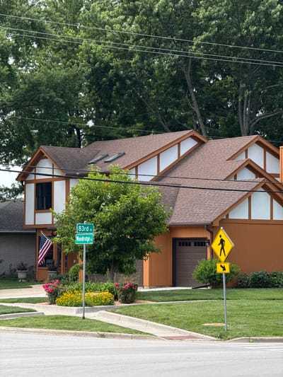 The image shows a residential street in Woodridge, United States of America. In the foreground, a paved road curves to the left, bordered by manicured green lawns and concrete curbs. Two street signs are visible: one green sign reads "83rd St" and "Woodridge Dr," indicating the intersection of these two streets. Above it, a yellow diamond-shaped sign depicts a pedestrian silhouette, a common "pedestrian crossing" warning sign. Below that, another yellow sign shows a black arrow pointing to the right.
A two-story house with a brown roof and distinctive orange siding dominates the midground. The house features white stucco panels with brown trim, and a dark brown garage door is visible on the right side. An American flag is displayed on the left side of the house. A mature tree with lush green foliage stands prominently in front of the house, partially obscuring its facade. Flower beds with pink and yellow blossoms are planted along the curb in front of the house.
In the background, a dense canopy of green trees forms a natural backdrop against a cloudy sky. Power lines are strung across the upper portion of the image, traversing in front of the trees. The overall scene depicts a well-maintained suburban environment.