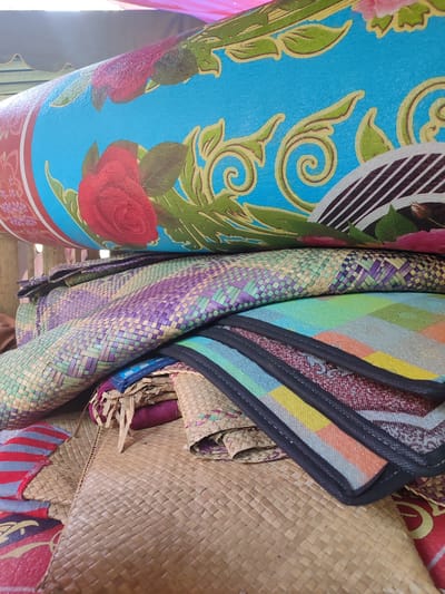 The image shows a stack of woven mats and a rolled-up mat with a floral design. The mats are made of natural fibers and some are woven with colorful threads. The rolled mat has a blue background with red roses and green leaves. The scene appears to be in a storage area, possibly a shop or warehouse, in Keudekarieng, Indonesia. The lighting suggests it is daytime, with light coming from above and to the left. No people are visible in the image.