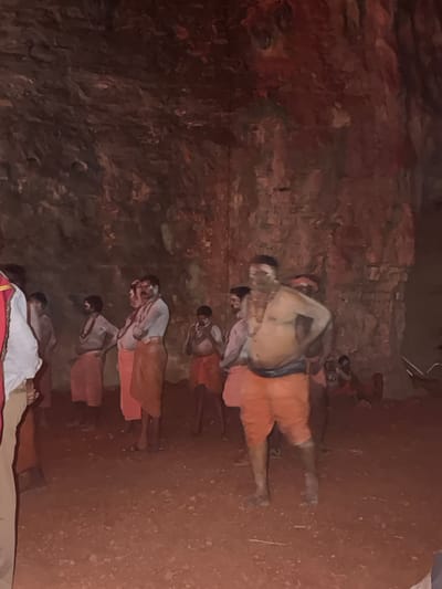 The image depicts a group of men dressed in traditional attire, possibly in a cave or cavern, given the rough, rocky walls and earthen floor. The men, primarily bare-chested with some wearing draped cloths around their waists, appear to be participating in a ritual or ceremony. Many have white markings on their faces and bodies, indicative of religious or cultural practices. The environment is dimly lit, with a reddish hue suggesting either natural light filtered through an opening or artificial lighting. The ground is covered in red soil and small stones. The context of the image places this scene in Bethamcherla, India, a location known for its caves and natural formations, suggesting this could be a religious site or a significant cultural gathering within such a natural setting.