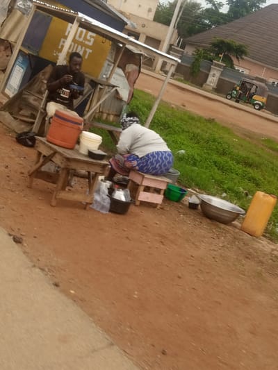 An outdoor scene depicts a roadside stall in Russo, Nigeria. A man, wearing a dark t-shirt and light trousers, is seated, eating from a blue plate. To his left is a large orange cooler and a white bucket on a wooden table. Behind him, a woman, dressed in a white top and patterned blue trousers, is seated on a small pink stool. Various containers, including a large silver basin, a green bowl, and a yellow jerrycan, are arranged on the ground near her. A three-wheeled vehicle is visible on the dirt road in the background, with a residential building and a fence to its right. The ground is reddish-brown dirt, and grass grows to the right of the stall.
