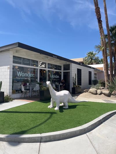 Here is a description of the image:
The photograph depicts the exterior of the "Wonder" restaurant, located at 74275 in Palm Desert, California. The building features white-tiled walls, glass windows displaying the restaurant's name and offerings (Breakfast, Lunch, Catering, Coffee, Beer, Wine, Cool Food Cool Drinks), and a black trim along the roofline. A large, white dinosaur statue stands on a patch of artificial turf in front of the restaurant. Palm trees are visible in the background against a clear blue sky. The surrounding area includes a concrete sidewalk and landscaping with rocks and plants. People are seated inside the restaurant.