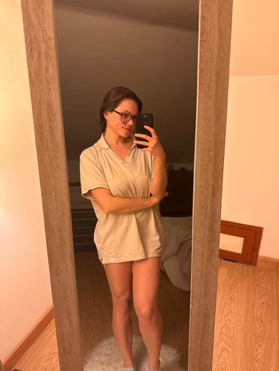 A full-length mirror selfie captures a woman standing indoors, presumed to be in a private residence in Cava de' Tirreni, Italy. The woman has dark hair and wears dark-rimmed eyeglasses. She is dressed in an oversized, light beige collared shirt, which extends to her upper thighs, revealing her bare legs. Her arms are crossed, with her right hand holding a black smartphone pointed at the mirror to record the image. She is looking towards the phone with a slight smile. The reflected environment features light wooden flooring, with a white, fluffy circular rug positioned beneath her feet. To the left of the woman's reflection, a portion of a bed with light-colored bedding is visible, and further back, a dark wooden dresser. To the right, a rectangular wooden frame rests against a light-colored wall on the floor. The scene is lit by warm, artificial indoor illumination.