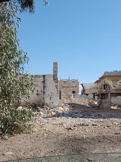 The image displays a scene of extensive urban destruction in Al-Qadam Municipality, Syria, under a clear blue sky. The foreground consists of barren, light brown earth covered in scattered small rocks and debris, with a dark horizontal line along the bottom suggesting the interior of a vehicle. A large, leafy green tree, possibly a eucalyptus, partially frames the left side of the scene.

The midground is dominated by widespread structural collapse, featuring substantial mounds of building rubble composed of shattered concrete, bricks, and plaster. Multiple heavily damaged buildings are visible. On the left, a light-colored structure exhibits numerous impact perforations across its remaining walls, with a tall, slender, vertical section, possibly a column or chimney, standing prominently amidst the debris. To the right, another multi-story, light-colored building shows significant damage, including pockmarked facades and missing sections that expose darkened interior spaces. A patterned concrete block parapet is visible on the roof of a structure in the upper-right midground. No people, animals, or active machinery are present, presenting a static depiction of a severely damaged urban environment.