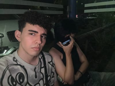 Two people pose closely for a photo. One wears a white t-shirt with a black graphic print and has curly hair. The other wears a black top and holds a phone, partially obscuring their face. They appear to be outdoors at night, with glimpses of a car and vegetation in the background. The image is rotated, giving a slightly disorienting perspective.