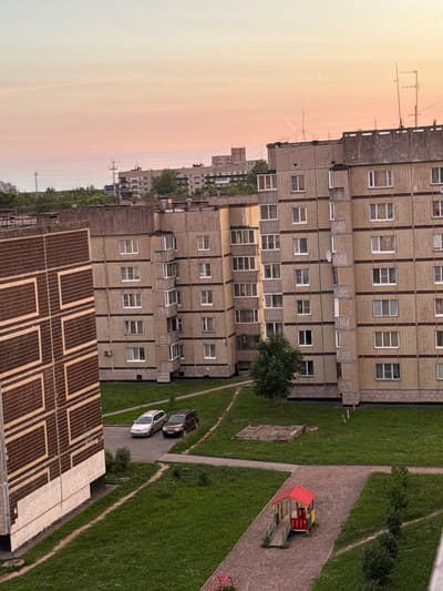 This is an aerial, long-shot, eye-level view of a residential courtyard in Kirishi, Russia, during what appears to be sunset. The sky is a soft gradient of pale yellow and pink hues. Dominating the foreground is a multi-story, beige brick apartment building with numerous windows and balconies. To the left, a section of another apartment building with a patterned brown and beige facade is visible.
In the courtyard, two cars are parked on a paved area: a silver station wagon and a dark brown SUV. A small, red-roofed playhouse with yellow and green accents sits on a paved pathway in the lower right quadrant of the image, accompanied by a small slide structure. A portion of a sandbox is also visible on the grass. Green grass covers the majority of the courtyard area. A few trees are scattered throughout the scene, including a young deciduous tree near the center of the image. The background features more apartment buildings and trees, with visible utility poles and antennas on the rooftops.