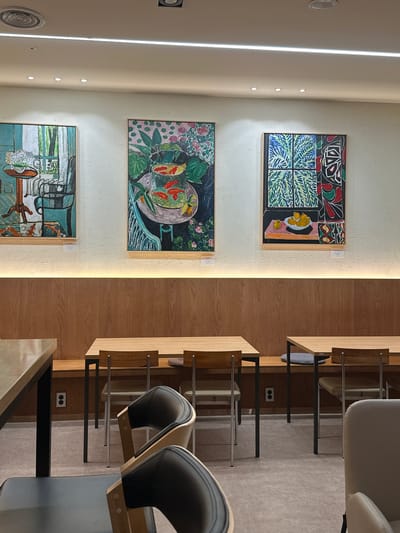 The image depicts an indoor scene, likely a cafe or restaurant, with artwork displayed on a textured white wall. Three framed paintings are prominently featured. The leftmost painting is a still life with a vase of flowers on a table, a chair, and a window view. The central painting is a still life of goldfish in a bowl with surrounding foliage. The rightmost painting features a still life of fruit on a table set against a patterned background. Below the artwork, a wooden wall paneling runs horizontally, illuminated by a strip of warm lighting. In the foreground, several tables and chairs are arranged. The tables are made of light-colored wood with dark metal legs. The chairs vary in design, with some featuring black upholstered seats and backs with wooden frames, and others appearing to be lighter upholstered chairs. The floor is covered with a light-colored, speckled material. Electrical outlets are visible on the wooden wall paneling. The scene appears to be set in Jeonju-si, South Korea, a city known for its cultural heritage and traditional architecture, although the interior design suggests a modern establishment. There are no people visible in the image.