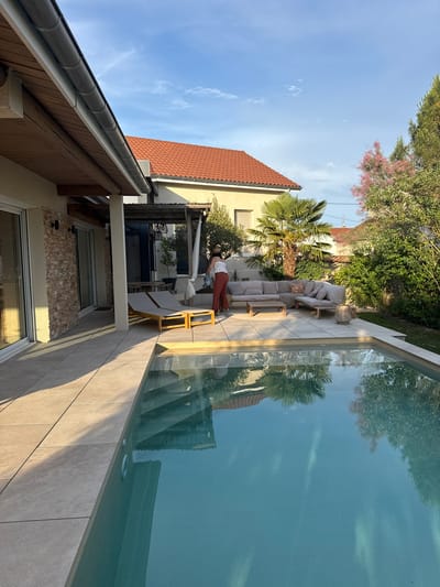 A woman wearing a white top and red pants is standing on a tiled patio next to a swimming pool in a backyard in Savigneux, France. She is holding a watering can and appears to be tending to a plant. The patio has two lounge chairs and a sectional sofa with throw pillows. A low coffee table sits in front of the sofa. In the background, there is a house with a red tiled roof, a palm tree, and various other plants and trees. The sky is blue with some scattered clouds. The swimming pool has clear blue water with reflections of the surrounding trees and buildings.