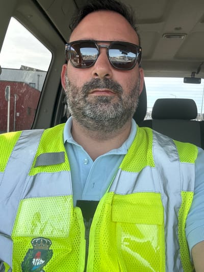 A man is wearing a hi-vis vest and sunglasses in the driver's seat of a vehicle. The vehicle appears to be in Wasserbillig, Luxembourg, as evidenced by the reflection in his glasses showing buildings in the town.