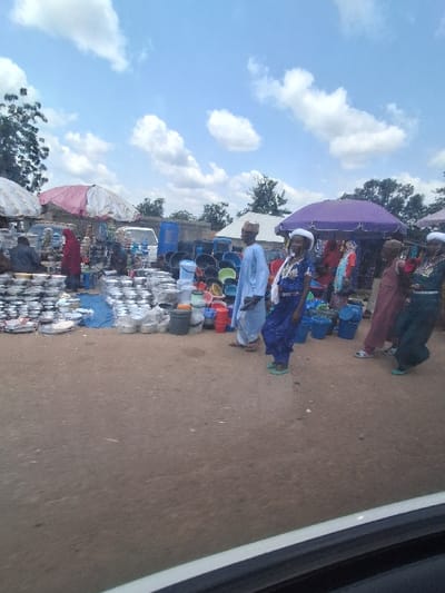 The scene depicts an active outdoor market situated on unpaved reddish-brown ground in Kungana, Nigeria, under a bright blue sky with scattered white cumulus clouds.
In the left foreground, the white frame of a vehicle is visible, indicating the vantage point. The market comprises multiple stalls, two of which are prominent: one under a light-colored umbrella (white or very pale pink) on the left, and another under a dark purple umbrella on the right.
The left stall features numerous stacks of various sizes of metal pots, bowls, and plates, likely aluminum or stainless steel, arranged directly on blue fabric and the ground. A person wearing a red garment and head covering is partially visible behind this stall. Further back, a white van is parked.
Towards the center and right, extensive displays of assorted plastic buckets and containers in colors including black, red, blue, and green are arranged on the ground.
Several individuals are present, dressed in traditional attire. In the mid-ground, a male wearing a light blue garment and a light-colored cap walks towards the right. To his left, a female in a dark blue patterned dress, white head covering, and light green footwear walks in the same general direction. Another female in a dark green garment and light green footwear is visible walking towards the right side of the frame. Multiple other individuals, predominantly female, wearing various colored dresses and head coverings (including red, maroon, and blue), are standing or moving near the purple umbrella stall and deeper into the market. The overall scene suggests ongoing commerce and pedestrian activity within the market environment.