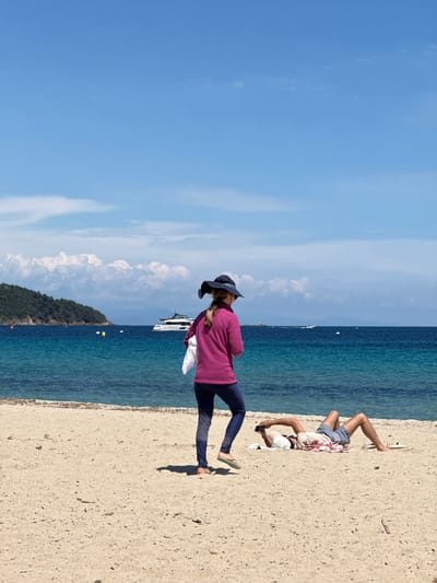 The image shows a beach scene in Ramatuelle, France. A woman wearing a purple long-sleeved shirt, dark leggings, a hat, and sandals stands on the beach. Beside her, a man is lying on the sand, partially obscured. The background features a blue sky, turquoise water, a small island with green foliage, and a yacht.