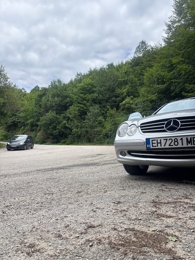 In the city of Shipkovo, Bulgaria, a silver Mercedes-Benz CLK-Class coupe is parked on a gravelly, winding road. The car, positioned in the foreground and angled towards the right, features a prominent license plate reading "EH 7281 MB" with a Bulgarian flag emblem. In the background, a dark-colored hatchback is visible further down the road. Both vehicles are situated amidst a lush, dense forest of green trees that flank the roadway, under a cloudy sky. The road surface appears to be a mix of asphalt and gravel, with scattered leaves and debris.