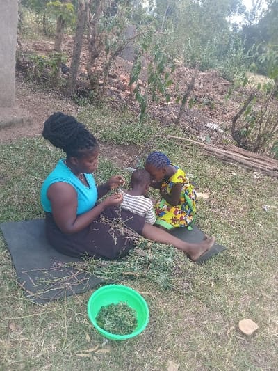 A woman with dark hair sits on a black mat in a grassy area, wearing a blue top and dark skirt. Two children, one in a colorful yellow dress and the other in a striped shirt, sit near her. They seem to be preparing food, as a green bucket filled with greens sits nearby. Debris, twigs, and grass surround them, and trees and vegetation form a backdrop. The scene suggests a rural or outdoor setting.