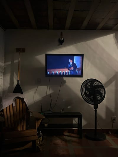 A wall-mounted TV displays a scene with a man in a suit. Above it is a black fan. A black metal shelf unit sits to the right, near a brick wall. Below the TV, a paddle is mounted horizontally to the wall, extending over a stack of wooden chairs covered with a white cloth. A wine bottle holder is on the left wall, with brick texture. The room appears to be dimly lit.