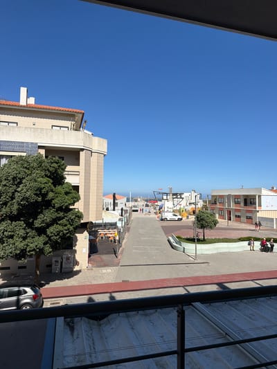 This is an outdoor, eye-level, medium shot of a street scene in Gafanha da Nazaré, Portugal, on a clear, sunny day. The foreground shows a black metal railing above a weathered, light-colored surface with parallel lines. The middle ground features a wide, paved walkway leading towards the horizon. On the left, a multi-story building with beige brickwork and a red-tiled roof is partially visible, with a large, dense green tree in front of it. A silver car is parked on the left side of the street. To the right of the walkway, there's a planter with a curved, light green bench where several people are seated, including a person in a wheelchair. Beyond the planter, a two-story building with pale orange and white facades is visible. The background shows more buildings, a glimpse of the ocean, and a clear blue sky. People are walking along the street and seated on benches, and vehicles, including a white van, are present. The overall scene depicts a public area with commercial buildings and pedestrian activity.