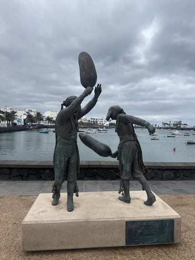 Two bronze statues of men throwing something, possibly bags filled with air, are prominently displayed against a waterfront backdrop. The statues are mounted on a concrete base, and a plaque is affixed to the side. The city of Arrecife, Spain, is visible across the water, featuring buildings, palm trees, and boats on the water. The sky is overcast with grey clouds.