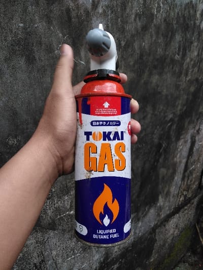 A light-skinned human hand is shown holding a "TOKAI GAS LIQUIFIED BUTANE FUEL" can vertically, positioned against a textured, dark gray wall. The cylindrical can is predominantly blue and white, featuring the brand name "TOKAI GAS" in orange letters and an orange flame logo. Japanese text, "日本テクノロジー," is visible on the upper white section. The top of the can is red, displaying visible red paint drips. A gray and white plastic torch head is attached to the red top. The background wall exhibits discoloration and weathering. There are no identifiable features within the image to determine a specific location in Mornington, Australia.