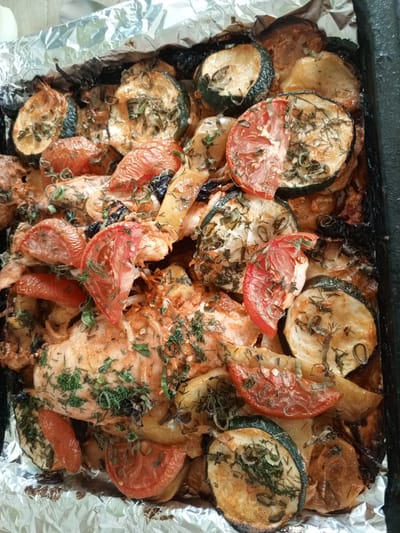 The image shows a close-up, overhead view of a rustic baked dish in a foil-lined baking pan. The dish contains pieces of chicken breast, sliced zucchini, sliced tomatoes, and sliced potatoes, all seasoned with herbs such as dill and what appear to be chili flakes. Some of the vegetables and chicken show signs of browning and caramelization from baking. The dish is quite full, with the ingredients piled together. The background shows a dark pan edge on the right and crumpled aluminum foil. The scene is consistent with a home-cooked meal. There is no identifiable information to suggest a specific location in Buraly, Russia.