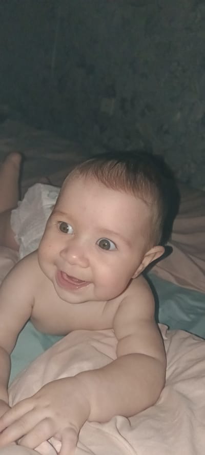This media file shows a close-up, low-angle shot of a baby lying on its stomach. The baby has light brown hair and is looking to the left of the frame with a wide-eyed expression and an open-mouthed smile, revealing its tongue and what appear to be gums or emerging teeth. The baby is wearing a white diaper.
The setting appears to be indoors, possibly a bedroom, with soft blankets or bedding in shades of pink and teal. The background is dark and out of focus, with some subtle textures visible that could suggest a wall or other soft furnishings.
The lighting is dim, creating a cozy atmosphere. The time of day is not explicitly indicated, but the low lighting could suggest evening or night. The baby's expression conveys happiness and engagement. There is no visible text in the image. The location context provided is "Хохлы, Russia."