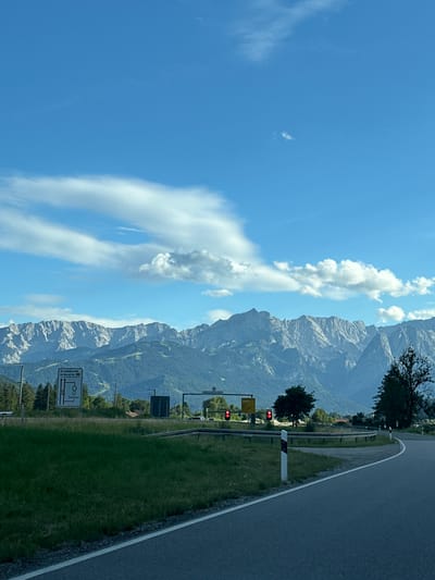 The image shows a road curving to the right in front of a lush green field with a backdrop of majestic mountains. The sky is a vibrant blue with scattered white clouds. Traffic lights are visible, indicating a road junction. A signpost with German text and symbols is present, which is related to road traffic and the town of Farchant. The overall scene depicts a serene landscape in Farchant, Germany.