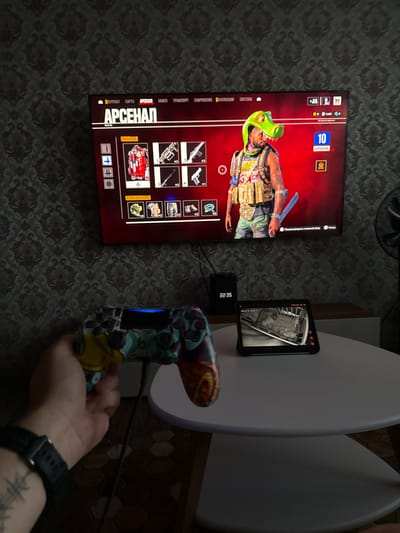 The image shows a gaming setup. A large television displays an in-game menu labeled "АРСЕНАЛ," which is Russian for "Arsenal." The menu features various weapons and character customization options. A person's hand is visible holding a video game controller, suggesting they are actively playing the game. On a small table in front of the TV, a tablet displays a black and white live feed, possibly from a security camera, showing a crib with what appears to be a baby. A small digital clock on the same table shows the time as 22:35. The background features a patterned wallpaper. The scene is indoors, likely in a living room, with a fan visible on the right side of the image. The presence of a gaming console, controller, and TV indicates a recreational activity. The tablet displaying a baby's room suggests a dual purpose for the setup, possibly monitoring a child while gaming. The image does not contain any information that would place it specifically in the city of Kondopoga, Russia.