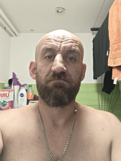 A man with a bald head, beard, and green eyes is looking at the camera in a bathroom. He is wearing a silver chain necklace. Behind him, there are several toiletries, including a box of detergent and a bottle of body lotion. There are also towels hanging on a rack, and a green tiled wall. The scene is lit by overhead lighting. The location is implied to be Stargard, Poland.