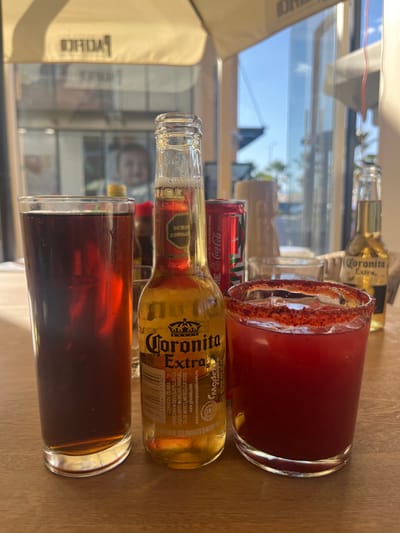 The image displays a close-up, eye-level perspective of three beverages positioned on a light-colored wooden table, suggesting a setting within an establishment in Santiago de Querétaro, Mexico. From left to right, the items are: a tall, clear glass filled approximately three-quarters with a dark brown, carbonated liquid; a 210 ml glass bottle of Coronita Extra beer, partially consumed, with its label clearly visible; and a short, wide clear glass containing a deep red liquid, likely a michelada, distinguished by a prominent red spice coating (chili powder or similar) on its rim and visible ice cubes. A red Coca-Cola can is partially visible behind the Coronita Extra bottle, and another Coronita Extra bottle is visible, out of focus, further to the right in the background. The background features large glass panels reflecting a bright blue sky, distant building structures, and a large, light-colored outdoor umbrella with reversed text, possibly "PACIFICO." A blurry reflection of a human face is also discernible in the upper left portion of the background reflections. The scene is brightly lit by natural daylight. No individuals are directly visible, nor are any specific actions or interactions depicted beyond the static arrangement of drinks.
