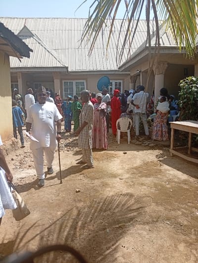 The image depicts an outdoor gathering of approximately 25-30 individuals in Unguwar Gaya, Nigeria, within what appears to be a residential compound. The ground is unpaved, consisting of dry, reddish-brown dirt, with shadows of palm fronds cast upon it in the foreground.
In the background, a light-colored building with a grey corrugated metal roof, white-framed windows, and decorative pillars on a porch or veranda is visible. A blue satellite dish is mounted on the building's exterior. To the left, a portion of another structure with a darker roof is present. On the right, a small green bush grows near the building, and a wooden table or bench is partially visible. A white plastic monobloc chair is positioned in the mid-ground, near the main building.
The central figure in the foreground is an adult male, of apparent African ethnicity, walking towards the viewer. He wears a white kaftan-style outfit, a patterned cap, and eyeglasses. He holds a dark walking stick in his right hand and a piece of white paper in his left. To his right, another male wears a light-colored, patterned tunic and trousers.
Most other individuals are congregated in the mid-ground and background, forming a casual group near the building's entrance. They consist of adults and some children, dressed in various traditional Nigerian attire, including colorful patterned dresses for women (e.g., green, pink, blue patterns) and tunics for men. One man stands out in a distinct red garment. A woman on the far right carries an infant secured to her back with a patterned cloth. Individuals appear to be standing, conversing, or observing. The scene is illuminated by bright daylight, casting distinct shadows.