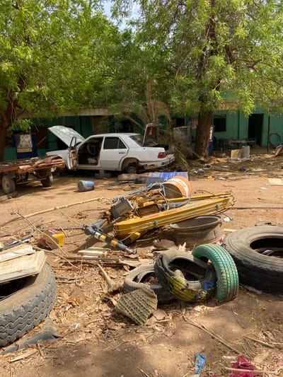 The image shows a scene of debris and destruction in Khartoum, Sudan, at the coordinates 15.552 latitude and 32.57 longitude. A white sedan with its hood open is parked on the dirt ground, surrounded by debris. A large, yellow, articulated crane arm is lying on its side, along with a variety of discarded tires. The tires are clustered in a pile, stacked on top of each other. The environment is dry and dusty, with visible remnants of leaves and branches scattered across the ground. The scene indicates a possible event of violence or destruction, highlighting the impact of unrest or conflict on the area.  A green building with windows and a doorway is visible in the background, partially obscured by foliage. The presence of the vehicle and crane suggests that the location may be a repair shop or a construction site.