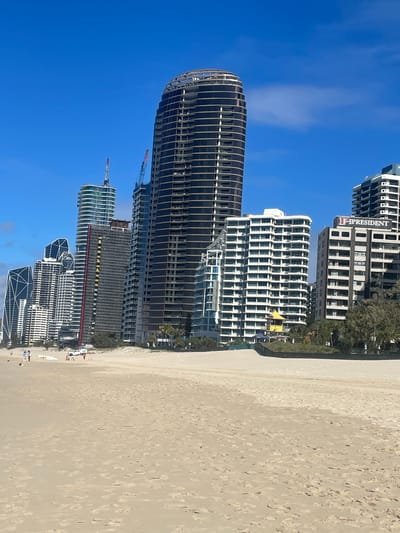 A wide shot captures a sunny day at a beach in the Gold Coast, Australia, with a clear blue sky and a few clouds. The foreground is dominated by a sandy beach. In the midground, a line of modern high-rise buildings forms the city skyline. Among them, a tall, dark, curved building stands out, possibly under construction with exposed floors and a skeleton-like top. Adjacent to it are several other apartment buildings with white facades and balconies. A construction crane is visible near one of the buildings. A sign reading "PRESIDENT" is mounted on one of the buildings. In the distance, palm trees and other greenery are visible between the buildings. A few people are scattered on the beach, some walking and others near a parked white vehicle and a surfboard. The ocean is not visible in this frame.