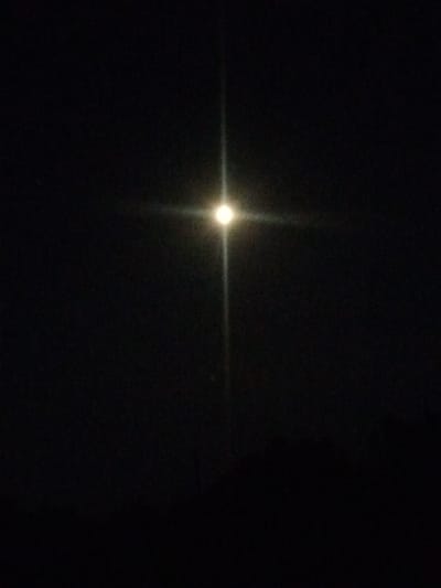 The image displays a dark, night sky with a bright, glowing object in the center, emitting a starburst effect. The object is likely the moon, heavily overexposed by the camera. Faint horizontal and vertical lines of light extend from the central glow, creating a cross-like appearance. The lower portion of the image shows a dark, indistinct horizon line, suggesting the presence of trees or buildings, but these are obscured by the darkness. The overall scene is characteristic of a nighttime photograph taken with a light source that is too bright for the camera's exposure settings. There are no people or discernible objects other than the celestial body and the faint silhouette of the landscape. The location is Kostiantynivka, Ukraine.