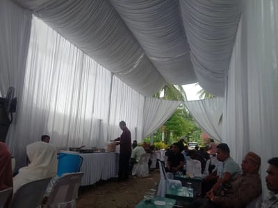 The image depicts an outdoor event, likely a reception or gathering, held under a white draped canopy. The canopy is supported by poles and has fabric panels creating a tent-like structure. The ground appears to be dirt or gravel.
In the foreground and midground, several people are seated and standing. On the left, a person wearing a white headscarf is seated. To their right, a table is set up with food, including serving dishes and containers. A man in a maroon shirt and black pants is standing at the table, appearing to serve food. Other individuals are seated at tables in the midground, some with white chair covers.
In the background, more people are visible, and the scene opens up to greenery, including palm trees, suggesting a tropical or subtropical environment. A white vehicle is parked in the distance. The overall atmosphere suggests a communal event.
The provided context indicates the location as Seuneubokpadang, Indonesia. This information helps to place the scene within a specific geographic and cultural setting, potentially influencing interpretations of the attire, food, and general ambiance.
