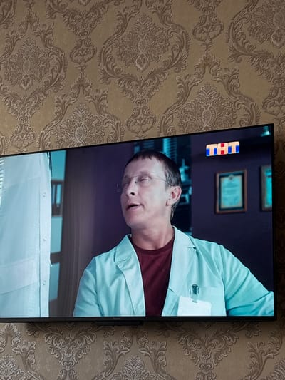 The media file shows a close-up shot of a television screen displaying a scene. The screen is oriented vertically. On the screen, a person wearing glasses and a light blue medical-style coat over a dark red shirt is visible. The person's head is tilted, and their mouth is slightly open, suggesting they might be speaking or reacting to something. To the left of the person, there's a stack of three colorful blocks, one red and two blue, forming a logo or symbol. In the background of the screen, there are framed pictures on a purple wall, and a glimpse of a table or desk with white surfaces. The television itself is black and is placed in front of a patterned wallpaper with an ornate floral design in shades of beige and brown. The overall impression of the scene on the TV is that it could be from a medical drama or a talk show. No specific time of day or weather conditions are discernible from the image. The location context provided is Nefteyugansk, Russia. The logo on the blocks appears to be a stylized "TNT" which is a Russian television channel.