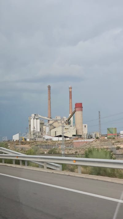 An industrial complex, likely a power plant, is visible in the distance under a cloudy sky. The facility features several tall smokestacks, some made of reddish-brown brick and others of lighter material. There are also large, industrial buildings with various structures, conveyor belts, and silos. A tall, cylindrical tank with a red top is prominent. Power lines and towers are present near the complex. In the foreground, a highway barrier and a section of the road are visible, suggesting the image was taken from a moving vehicle. The landscape around the industrial site is dry and arid, with sparse vegetation. While the exact location in Zaragoza, Spain is not specified, this type of industrial facility is commonly found on the outskirts of urban areas. No people or specific actions are depicted.