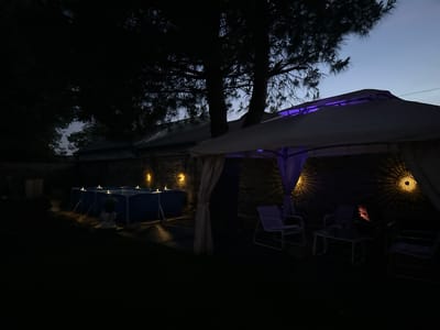 This image depicts an outdoor scene at dusk or night, likely in the garden of a property in Villa Pasquali, Italy. The primary focus is on a backyard area, featuring a temporary above-ground swimming pool, a gazebo, and patio furniture.

The swimming pool, a blue rectangular structure, is located on the left side of the frame and is illuminated by several small, strategically placed lights. The water's surface is calm and reflects the surrounding artificial lights.

To the right of the pool is a gazebo, its white canopy offering shade. The gazebo is adorned with purple LED lighting, casting a subtle glow. Underneath the gazebo, several chairs and a small table are arranged, suggesting a relaxation or gathering space. One of the chairs is occupied by a person who is illuminated by the light of a mobile device they are holding. This person is partially obscured by darkness, with only their face and hand visible.

The background consists of a stone wall, likely part of the property's perimeter, and silhouetted trees. Decorative wall lights with intricate patterns are mounted on the wall, adding to the ambiance. The sky above is a deep twilight blue. The overall impression is that of a tranquil evening setting, possibly after sunset, with ambient lighting creating a cozy atmosphere.