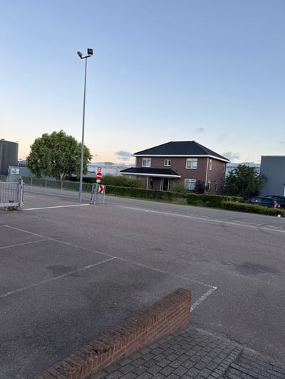 The image depicts an outdoor scene in the city of Maasdijk, Netherlands. In the foreground, a paved area, likely a parking lot, is marked with white lines. A low brick wall runs along the edge of the paved area, transitioning to a brick pathway. In the middle ground, a metal fence separates the paved area from a road and a residential area. A street lamp stands prominently on the left side of the image. Several traffic signs are visible, including a speed limit sign of 30 and a no-entry sign. A red and white directional sign indicates a turn to the right. Beyond the fence, a two-story brick house with a dark tiled roof is situated. Several windows are visible on the facade of the house. A neatly trimmed hedge surrounds the house. In the background, a large industrial building with a grey facade is visible, along with a tall green tree. The sky is clear and light blue, suggesting it is either early morning or late afternoon. There are no people visible in the image.