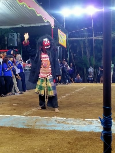 The image depicts a nighttime outdoor performance in Tamansari, Indonesia, taking place on a dirt ground marked with faint white and blue painted lines. A central barefoot performer, positioned slightly right of center, stands with their right arm raised, palm open. This individual wears a red and white mask with stylized features, a dark shaggy wig, a black long-sleeved jacket, a horizontally striped t-shirt featuring the word "PONOROGO" and a tiger graphic, and a green and yellow plaid sarong worn over dark trousers.
To the left of the performer, a group of spectators, including individuals in blue shirts and some wearing hijabs, observes the scene. They are situated under a white tent structure with decorative red and green trim. A large grey speaker is visible beneath the tent. To the right and in the background, more spectators are present, with some seated directly on the ground. The far background consists of dark, indistinct foliage, likely trees. The entire scene is brightly illuminated by multiple artificial lights, which produce prominent lens flares in the upper center and right of the frame. A dark vertical pole, wrapped with blue rope, is present in the extreme foreground on the right side. A yellow banner with black text is visible in the mid-ground right, partially obscured by the tent structure.