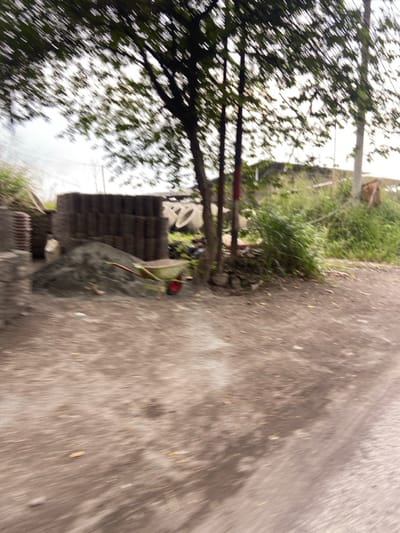 The image shows a dirt road in Kecamatan Margaasih, Indonesia, at the coordinates -6.961 latitude and 107.536 longitude. A large tree is in the foreground, with a pile of gravel and a wheelbarrow to the left. Behind them is a stack of cinder blocks and a few other large, circular objects. The image is taken from a moving vehicle, giving it a blurry appearance. The road continues into the distance, where there are trees and foliage on both sides. There are no people visible in the image.