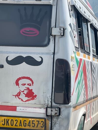 The image shows the rear and side of a white bus, likely in Ujroo, India, as indicated by the license plate "JK02AG4573". The bus has several decals. On the rear window, there is a dark gray silhouette of a crown with a pink oval shape within its band. Below the window, a black silhouette of a handlebar mustache is centered. Beneath the mustache, a red stencil of a man's face is depicted, with him appearing to hold a cigarette. A reflective red and white strip is visible above the license plate. The side of the bus features a series of colorful diagonal stripes in red, green, and white, along with some text that is partially obscured but appears to include "JK02AG 4573". The bus shows signs of wear, with peeling paint and rust on the hinges. The background suggests an outdoor setting, with a hint of mountains in the distance.