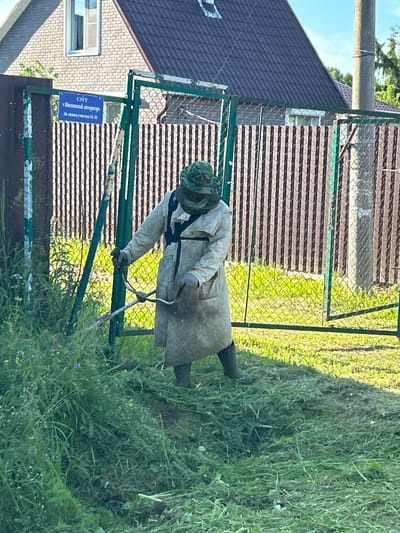 A person wearing a protective suit and a camouflaged hood is operating a brush cutter to mow tall grass. The person is standing near a green metal fence with a sign that reads "СНТ «Московский Инспектор» Заявки участки 71-32". Behind the fence is a house with a dark tiled roof and a brick facade. The scene appears to be in a suburban or rural area, possibly a dacha or garden community, given the presence of the fence and the sign. The ground is covered with cut grass, indicating recent activity. The sky is blue with some clouds.