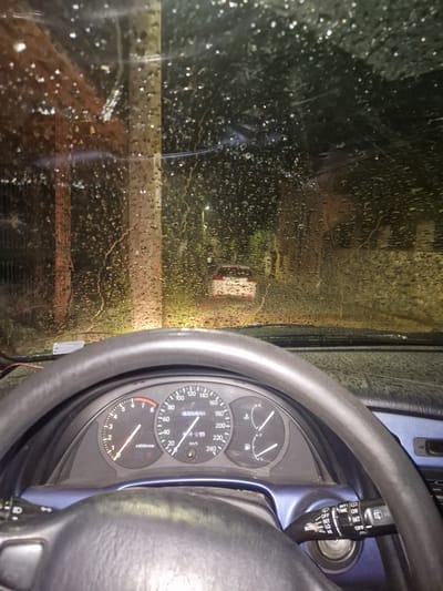 The image captures an interior view from a car, likely in Rila, Bulgaria, at night or in very low light conditions, during or immediately after rainfall. The primary subject is a car's dashboard and a rain-soaked windshield.

On the right side of the frame, the black steering wheel and a section of the dashboard are prominent. The instrument cluster features a speedometer marked up to 240 km/h and a tachometer reaching 8 x1000r/min, both indicating zero, suggesting the car is stationary or off. A digital display shows "325145.5" and "460," likely representing an odometer and trip meter respectively. Fuel and temperature gauges are also visible. To the right of the steering wheel, a wiper control stalk is seen, featuring various settings such as "INT" (intermittent), "OFF", "ON", "HI" (high speed), "LO" (low speed), "MIN", "TIME", and "MIST," along with corresponding icons. The car's interior trim around the dashboard and steering column appears blue.

The left side of the image is dominated by the wet windshield, covered with countless individual raindrops and streaks of water. Through the blurred, rain-distorted glass, a dark, indistinct exterior scene is visible. A bright, yellowish light source in the distance casts a significant glow, indicating the presence of artificial illumination like streetlights or another vehicle's headlights. Other fainter, diffused lights contribute to the dark, wet ambiance outside the vehicle. The scene conveys a sense of being sheltered inside the car while observing a rainy environment.