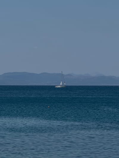 A sailboat is sailing on a calm, blue sea under a clear sky. In the background, a range of hazy blue mountains forms the horizon. The sailboat, with its white sails, is positioned slightly to the left of the center of the image. The sea is a deep blue, with gentle ripples. There is a small, orange buoy visible in the water in the lower left quadrant of the image. The scene appears to be set in Alimos, Greece, given the clear sky and the type of landscape depicted.