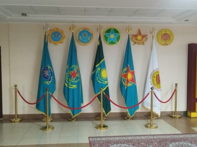 A well-lit indoor scene shows a row of seven flags displayed against a plain, cream-colored wall. The flags are mounted on poles with decorative gold finials at the top. They are arranged with red velvet ropes on gold stanchions in front of them, creating a barrier. Each flag is a different design, with prominent colors including teal, blue, green, yellow, and white. Some flags feature emblems or symbols, such as stars, suns, and circular designs with text. Above the flags, several circular emblems are affixed to the wall. These emblems are colorful and detailed, featuring more stars, suns, and what appear to be stylized wings or branches. The floor is tiled with a light beige pattern, and a section of a patterned rug is visible in the foreground. The overall impression is of a formal display, likely in a government building or public institution. The context of Қотырбұлақ, Kazakhstan, suggests this could be a governmental or ceremonial space within that region.