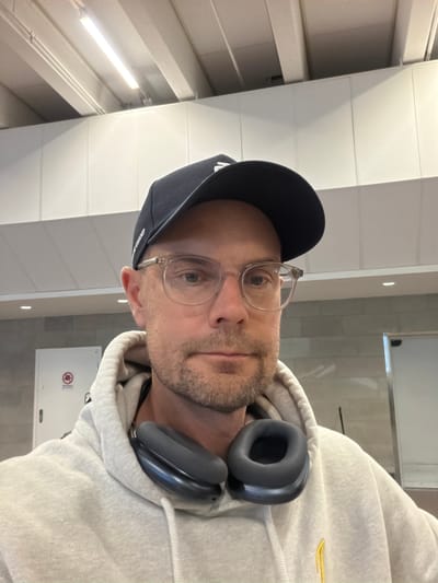 A man wearing a light grey hoodie, a black baseball cap, and clear-framed glasses is posing for a selfie. He has headphones draped around his neck and a light beard. The background suggests an indoor environment, possibly an airport terminal, with white paneling on the walls and ceiling, and fluorescent lighting. A closed white door with a red circular sign is visible on the left side of the frame. The man is looking directly at the camera. The scene appears to be a candid shot taken by the man himself.