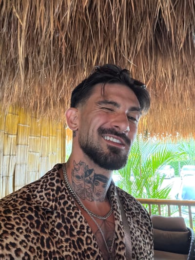 A man with dark hair and a beard is smiling and winking at the camera. He is wearing a leopard-print shirt and a silver chain. He has tattoos on his neck. The background is a thatched roof and bamboo wall. Palm trees and what appears to be a vehicle can be seen through an opening. The photo was likely taken in a shaded area. It could be in the city of Ban Khlong Khian บ้านคลองเคียน, Thailand.