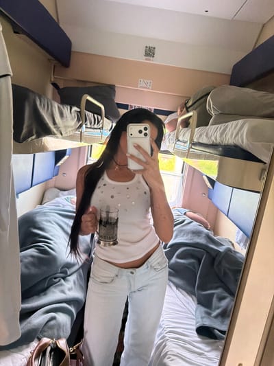A woman is taking a mirror selfie in a train compartment. She is holding a phone in her right hand and a glass with a metal holder in her left hand. She is wearing a white tank top with sequins and light-colored pants. The train compartment has bunk beds with blue and dark gray bedding. Another person is visible lying on an upper bunk in the background. The scene appears to be set within a train traveling through a rural or suburban landscape, indicated by greenery visible through the window. The location is identified as Babyakovo, Russia.