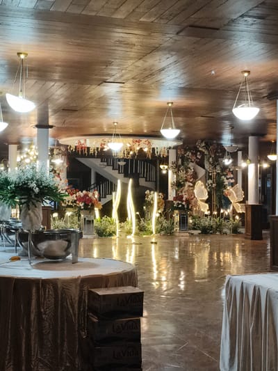 This image depicts an elaborately decorated indoor event space in Kawungluwuk, Indonesia, prepared for a formal gathering. The high ceiling is constructed from horizontal wooden planks, from which multiple brass-colored pendant lights with inverted bowl-shaped white shades are suspended, illuminating the area. The floor is highly reflective, likely polished tile or stone, showing distinct reflections of the overhead lights and various decorative elements.
In the left foreground, a round table draped with a wrinkled, dark beige tablecloth stands, with a stack of cardboard boxes labeled "La Vida" placed beneath it. To the right of this table, another table features a large chafing dish setup, positioned next to a tall, textured grey vase holding a substantial arrangement of white and green foliage.
The mid-ground and background showcase extensive floral decorations and lighting. A prominent black-railed staircase ascends on the left side of the background. Below and around the staircase, and extending across the mid-ground, are numerous floral arrangements in varying colors, including white, red, and orange, set on pedestals and within ornate structures. Several tall, narrow, illuminated light fixtures resembling abstract flames or pillars are visible, interspersed with foliage. On the right side of the background, large, illuminated butterfly-shaped decorations are incorporated into an archway adorned with more flowers and greenery. Additional smaller pendant lights and chandeliers are visible further back. No individuals are present in the scene.