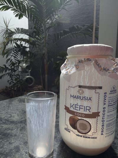 The image showcases a bottle of Marusia Tradicion Rusa Kefir Coco, positioned near a partially filled glass. The bottle's label prominently displays "Kefir Coco" with an image of a coconut. Text on the label details the probiotic nature of the drink and suggests shaking before opening. The glass contains a white, liquid substance, presumably the kefir. A plant is visible in the background, creating a natural setting. The composition is set against a muted gray surface.