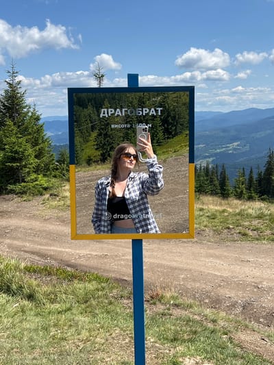 In Chorna Tysa, Ukraine, a young woman with braided brown hair, wearing sunglasses, a plaid shirt over a black top, and denim shorts, takes a selfie in a reflective sign. The sign displays the Cyrillic text "ДРАГОБРАТ" and "висота 1400 м," indicating "Dragobrat" and "height 1400 m." The sign has a blue pole and a yellow frame. The background reveals a mountainous landscape with lush green pine trees, rolling hills, and a clear blue sky with white clouds. The foreground shows a dirt path and grassy terrain. The woman is holding a smartphone to capture her reflection.