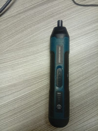 A black and teal cordless screwdriver with a dark gray head rests vertically on a light gray, wood-patterned surface. The screwdriver has the text "CORDLESS SCREWDRIVER" printed vertically on its body. Below this text are two buttons, one with upward and downward arrows and a central button. Further down, there is a battery indicator with the word "MAX" at the top and a symbol of a lighthouse enclosed in a circle. The background is a plain wood-grain texture. The image appears to be a product photo or a personal item on display, and there is no indication of the city of Shiroro, Nigeria, or any contextual information to place it there.